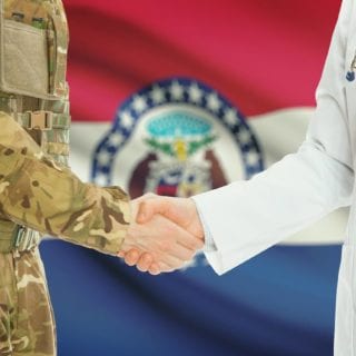Soldier in uniform shaking hands with doctor with Missouri state flag in background