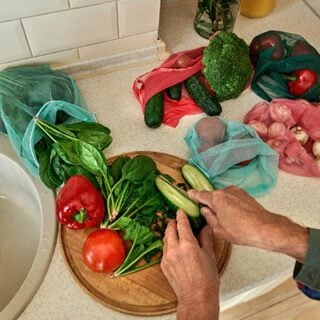 A pair of hands slicing a cucumber next to a sink. Other fresh produce is around the hands, including red peppers, cucumbers, tomatoes, mushrooms, basil and broccoli.