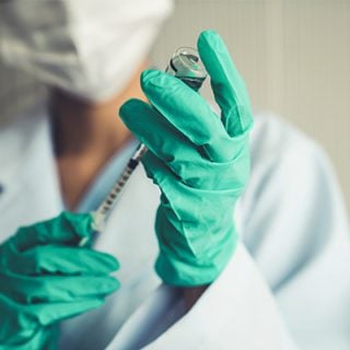 A masked doctor wearing green gloves uses a small syringe to withdraw an experimental cancer vaccine from a glass vial. The vaccine will be part of a mesothelioma treatment regimen.