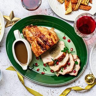 A holiday meal featuring sliced turkey on a green plate with gravy in a white gravy boat. A bowl of mashed potatoes, a plate of roast vegetables, a glass of water and glasses of red wine are also seen. Gold ribbons and stars decorate the place setting.