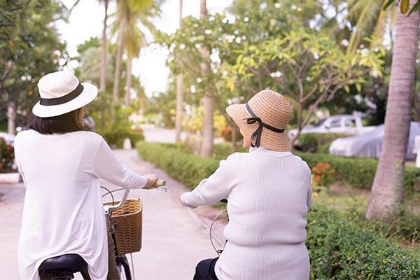 A mesothelioma patient and her friend are wearing sun hats and recreationally riding beach cruiser bicycles together.