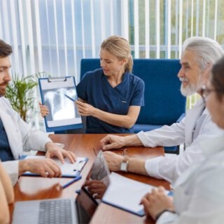 A multidisciplinary tumor board sits at a rectangular table reviewing a mesothelioma case. The board includes a thoracic surgeon, a radiologist, a pathologist and a medical oncologist.