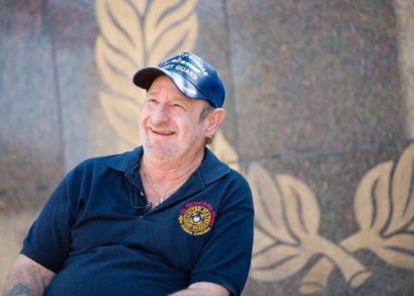 This photo shows a smiling Coast Guard veteran wearing a blue Coast Guard hat and polo shirt. He is sitting in front of a monument. Coast Guard veterans may have been exposed to asbestos during service.